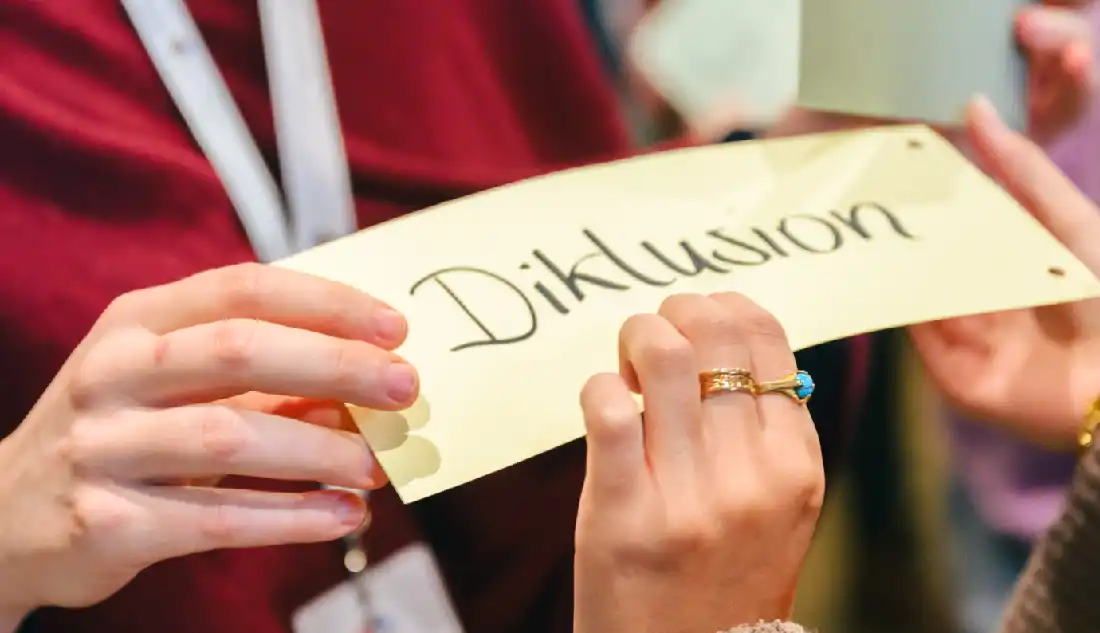 Zwei Hände halten ein Kärtchen mit der handgeschriebenen Aufschrift „Diklusion“.