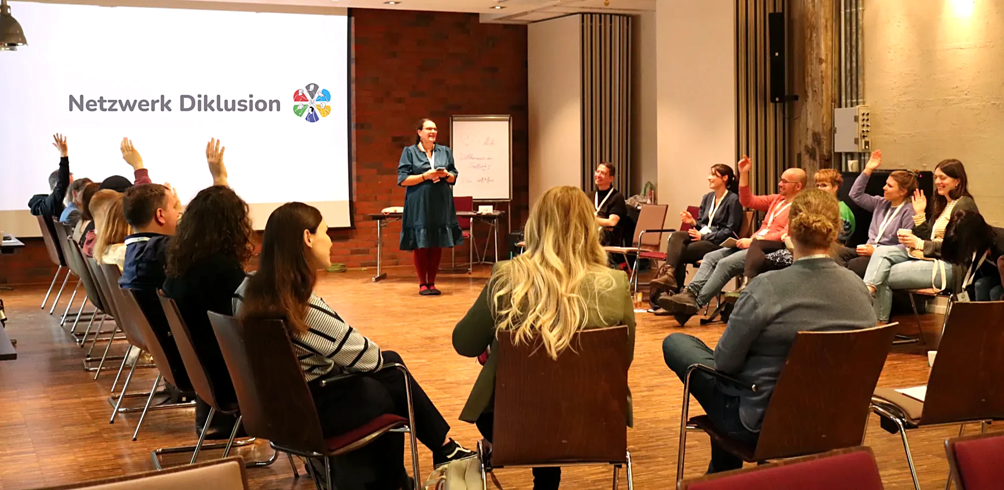 Dr. Lea Schulz bei einem Netzwerktreffen im Austausch mit Teilnehmenden, die sich mit erhobenen Händen beteiligen. Im Hintergrund die Leinwand mit dem Logo „Netzwerk Diklusion“.