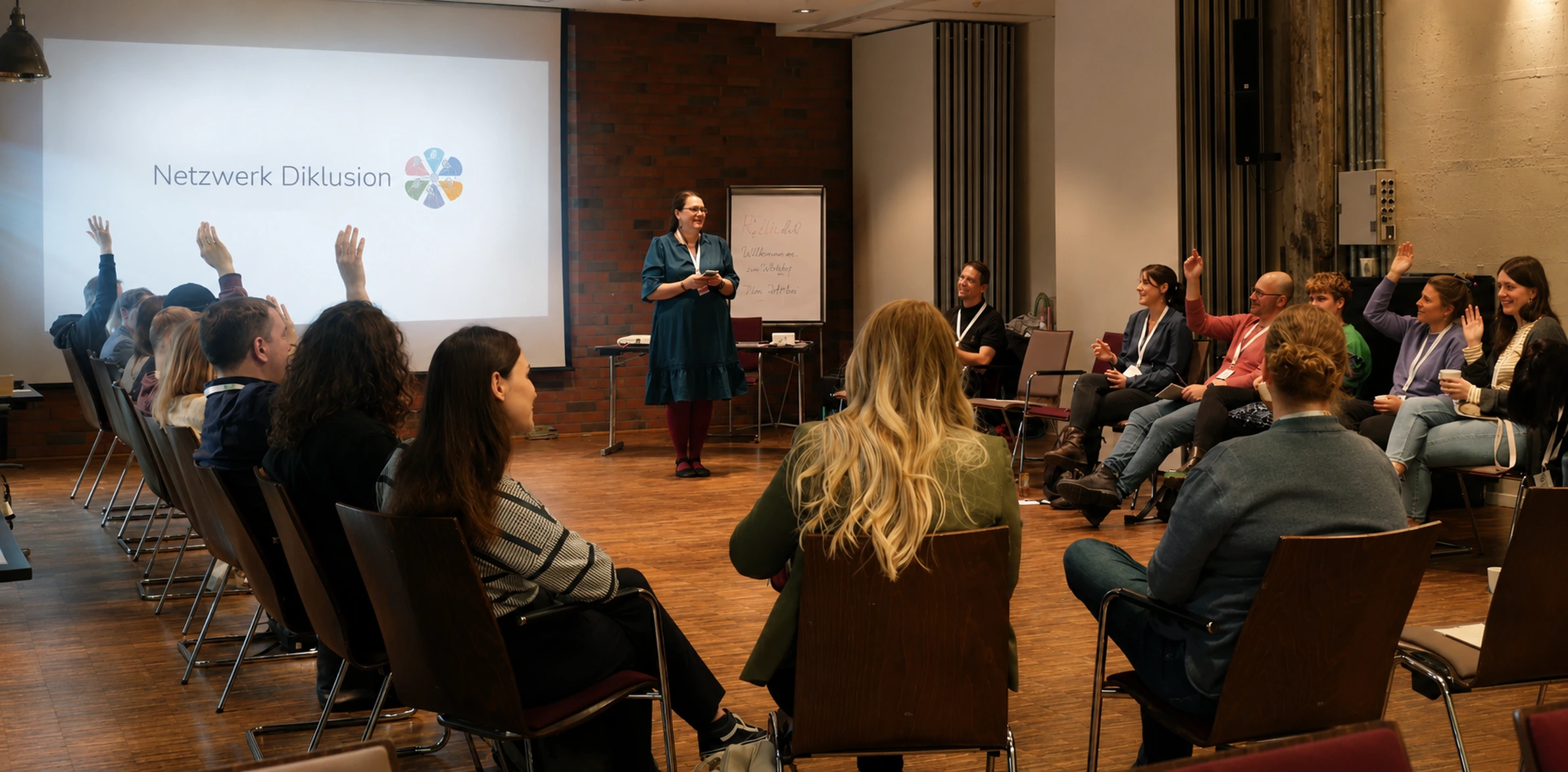 Dr. Lea Schulz bei einem Netzwerktreffen im Austausch mit Teilnehmenden, die sich mit erhobenen Händen beteiligen. Im Hintergrund die Leinwand mit dem Logo „Netzwerk Diklusion“.